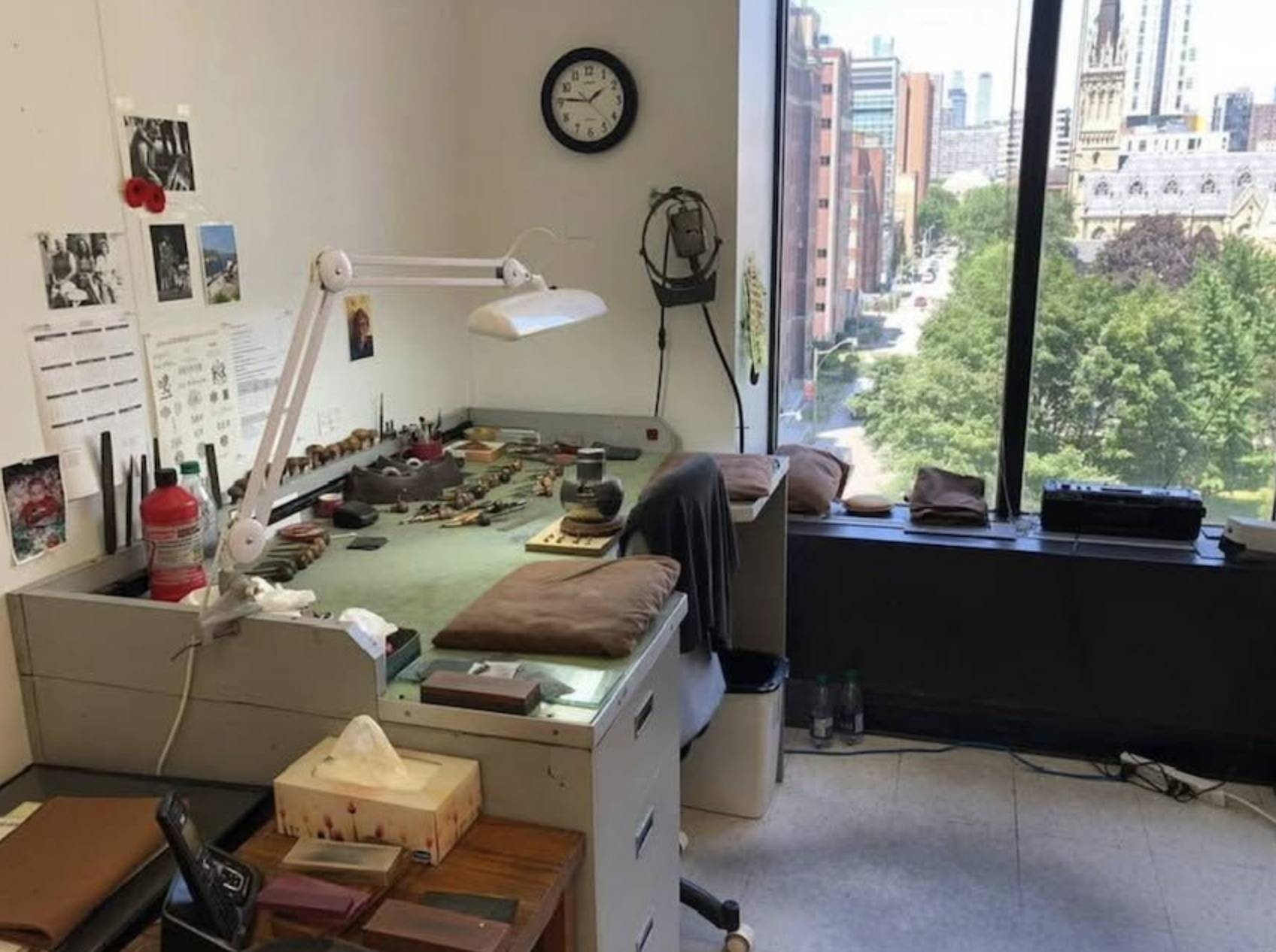 A hand engraver's workbench with tools, lamp and leather pads beside a window overlooking downtown Toronto.
